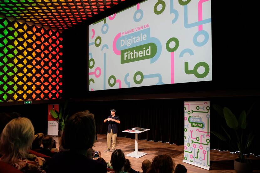 Dagvoorzitter verzorgt vanaf podium in grote zaal de opening van de Maand van de Digitale Fitheid