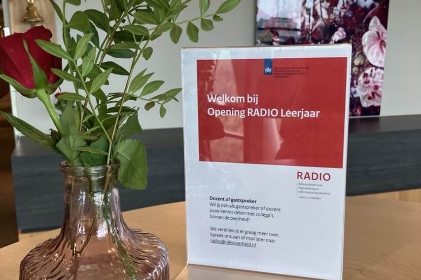Vaasje met rode roos naast staander met tekst 'Welkom bij opening RADIO Leerjaar'