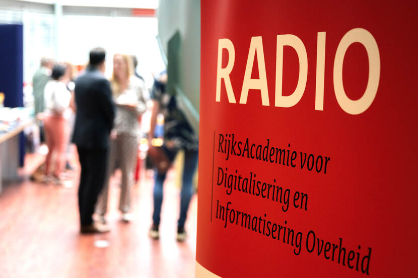 Banner op voorgrond met tekst 'RADIO RijksAcademie voor Digitalisering en Informatisering Overheid' met achtergrond aanwezigen bijeenkomst